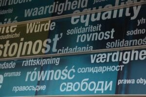 Na szklanej fasadzie budynku widoczne są duże napisy w różnych językach, przedstawiające słowa związane z wartościami takimi jak godność, równość i solidarność.